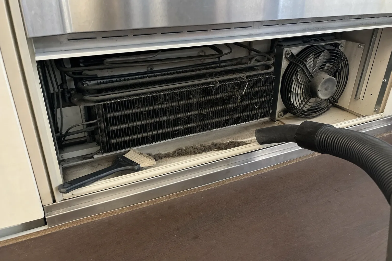 Condenser coil matted with dust and lint behind a removed base grille, with a brush and vacuum hose alongside