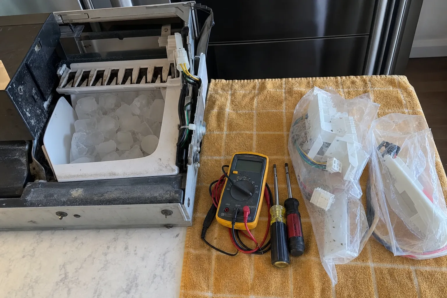 Ice maker module and its full bin set out on a counter beside a multimeter, screwdrivers and bagged parts